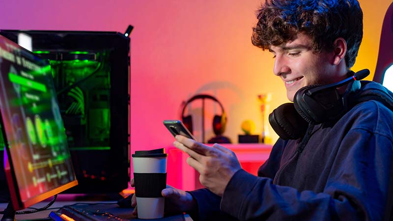 Imagem exibe jovem rapaz sorridente, bebendo café e utilizando smartphone. 